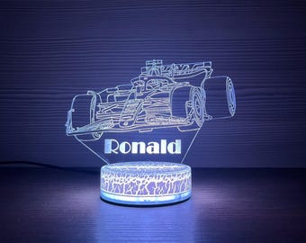 May include: A neon-lit, 3D-style Formula 1 race car lamp. The lamp features a detailed outline of a race car with the name "Ronald" beneath it. The base has a cracked pattern design, and the lamp emits a blue glow.