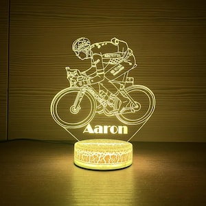 Może przedstawiać: Żółto podświetlana lampa akrylowa przedstawiająca kolarza na rowerze, z imieniem "Aaron" u podstawy. Lampa ma okrągłą podstawę z pękniętym wzorem. Kolarz ma na sobie kask i strój kolarski.