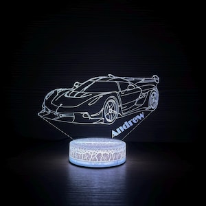 May include: A white LED night light featuring a sports car design. The car outline is illuminated, with the name "Andrew" displayed on the base. The base has a cracked pattern and emits a soft glow, creating a decorative and functional piece.