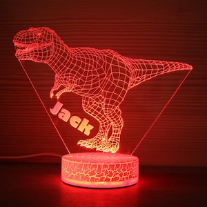 Puede incluir: Luz de noche de dinosaurio 3D roja con el nombre "Jack" mostrado. La figura del T-Rex está iluminada con un patrón de cuadrícula, y la base tiene una textura agrietada. La luz emite un brillo rojo cálido, creando una pieza decorativa única.