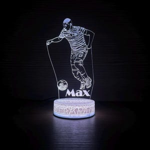 以下が含まれることがあります： ストライプのジャージを着たサッカー選手が特徴の、光るLEDランプ。ベースには「Max」という名前が書かれています。 ランプは、テクスチャ加工が施された丸いベースで、柔らかい白色光を発します。 サッカー選手はダイナミックなポーズをとっています。