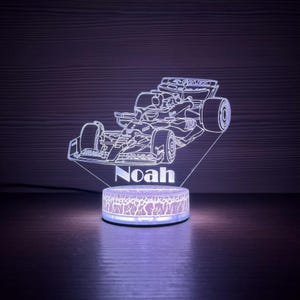 May include: A Formula 1 race car illuminated night light with the name "Noah" displayed. The light has a white outline of the car and a patterned base that glows with a purple hue. The background is dark.
