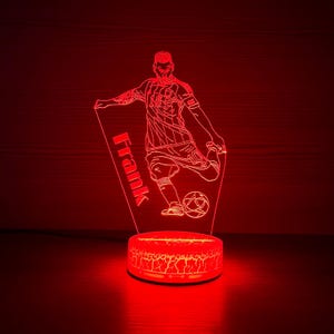 Puede incluir: Lámpara LED roja que representa a un jugador de fútbol pateando un balón. El jugador está representado en una pose dinámica, con el nombre "Frank" impreso verticalmente. La lámpara tiene una base con efecto de tierra agrietada.