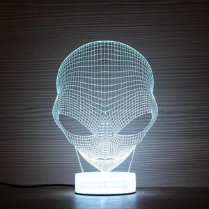 May include: A glowing, 3D-effect alien head lamp. The lamp features a grid-like design, with a white base and a light blue glow emanating from the alien's head. The lamp is a unique decorative item.
