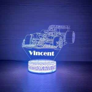 Könnte beinhalten: Ein Nachtlicht mit einem Formel-1-Rennwagen und dem Namen "Vincent" in Weiß beleuchtet. Das Licht hat einen kreisförmigen Sockel mit einem Muster. Das Farbschema ist blau und weiß.