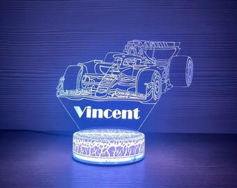May include: A Formula 1 race car night light with the name "Vincent" illuminated in white. The light has a circular base with a patterned design. The overall color scheme is blue and white.