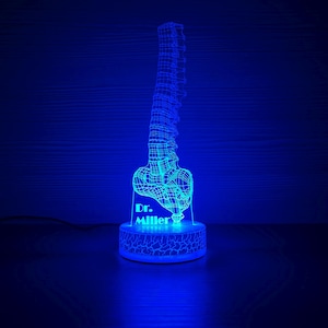 Könnte beinhalten: Blau beleuchtete 3D-gedruckte Wirbelsäulen-Nachtlampe. Der Sockel der Lampe hat ein Rissmuster und den Text "Dr. Miller". Die Wirbelsäule ist ein detailliertes Drahtgitterdesign. Das Licht steht auf einer dunklen Oberfläche.
