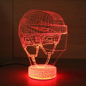 以下が含まれることがあります： 赤色の3D効果ホッケーヘルメットのナイトライト。ヘルメットはワイヤーフレームデザインで、透明なバイザーとあご紐が付いています。ベースは丸く、テクスチャ加工されており、温かい光を発しています。