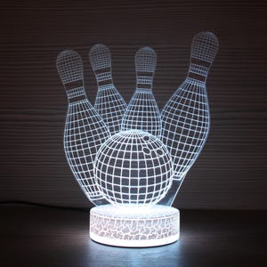 May include: A white LED lamp in the shape of a bowling ball and pins. The lamp has a grid-like design, with the ball in front of four pins. The round base of the lamp is also illuminated, creating a glowing effect.