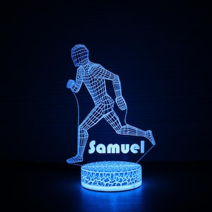 Puede incluir: Luz nocturna LED azul con un diseño de figura corriendo en forma de alambre. El nombre "Samuel" se muestra en blanco. La base de la luz tiene un diseño de patrón agrietado. La luz está sobre una superficie oscura.