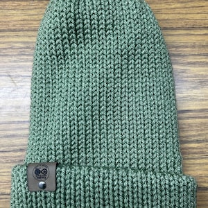 May include: A sage green knit beanie hat with a folded brim. The hat has a ribbed texture and a small, brown leather tag with a logo and text. The hat is made of a soft, warm material, perfect for cold weather.