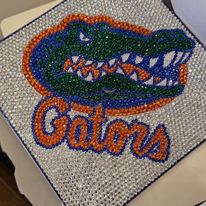 Könnte beinhalten: Eine Abschlusskappe, verziert mit glitzernden Strasssteinen. Das Design zeigt das Wort "Gators" in Orange und ein grünes und blaues Alligator-Logo. Die Kappe hat einen marineblauen Rand.