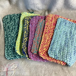 Könnte beinhalten: Eine Sammlung von sechs handgehäkelten Waschlappen in verschiedenen Farben. Die Waschlappen sind grün, gelb mit blau, lila mit türkis, rosa mit gelb und blau mit grün. Sie sind auf einer weichen, grauen Oberfläche angeordnet.