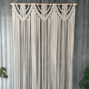 Può includere: Appeso a parete in macramè color crema. La parte superiore presenta un motivo geometrico decorativo, con lunghi fili verticali di cotone annodato. Il macramè è sospeso a un tassello di legno naturale, creando un elemento di arredo per la casa in stile bohémien.