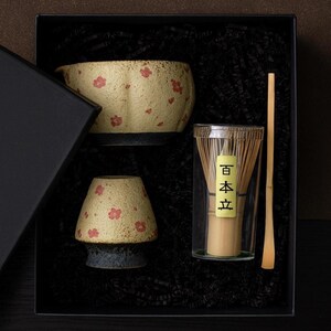 May include: A matcha tea set in a black gift box. The set includes a ceramic bowl and a smaller ceramic piece, both with a beige background and red floral patterns. A bamboo whisk and scoop are also included.