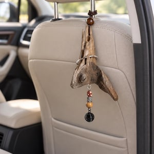 May include: A hanging car ornament featuring a piece of weathered driftwood suspended by a dark brown leather cord. The driftwood is accented with a silver metal ring and a dangling charm composed of patterned beads and a small black metal cage bead.
