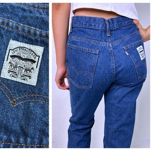 May include: Medium wash blue denim jeans featuring a high-waisted fit, straight legs, and a white Levi Strauss & Co. patch on the back pocket. The jeans include classic orange contrast stitching, a button fly, and a small white tab on the pocket.