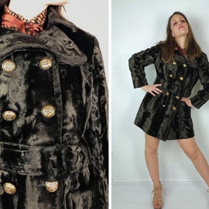 May include: A dark green velvet coat with gold-colored buttons and a matching belt. The coat has a double-breasted design and a notched collar. The buttons are ornate, adding a touch of elegance to the garment. The coat is knee-length.