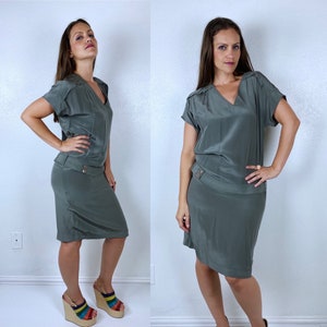 May include: A sage green silk dress with a v-neck and short sleeves. The dress has a skirt that falls to the knee and a fitted bodice. The dress is shown on a model.