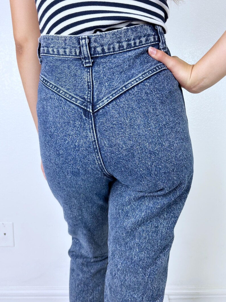 May include: Acid wash denim jeans with a high waist and a unique V-shaped yoke detail. The jeans have a classic five-pocket design and a straight leg. The denim is a medium blue wash with a slightly faded appearance, giving them a vintage look.