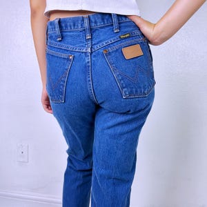 May include: Medium blue denim jeans featuring a high-waisted fit, a classic five-pocket design, and a tan leather patch on the back pocket with the Wrangler brand name. The back pockets include a signature W-shaped stitching detail in dark thread.