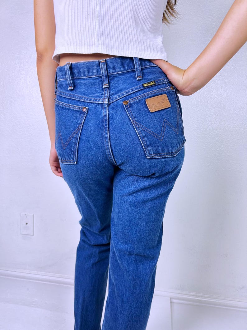 May include: Medium blue denim jeans featuring a high-waisted fit, a classic five-pocket design, and a tan leather patch on the back pocket with the Wrangler brand name. The back pockets include a signature W-shaped stitching detail in dark thread.