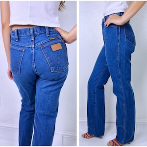 May include: Medium blue denim jeans featuring a high-waisted fit, straight legs, and a classic five-pocket design. The back pockets display a signature W-shaped stitching pattern in brown thread and a tan leather-like patch with the Wrangler logo.