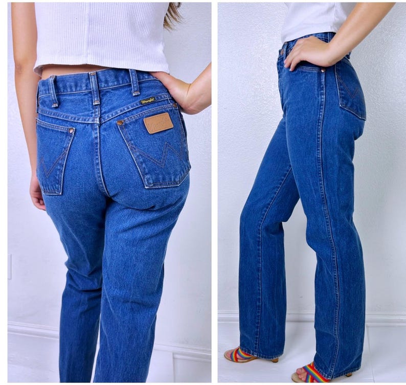 May include: Medium blue denim jeans featuring a high-waisted fit, straight legs, and a classic five-pocket design. The back pockets display a signature W-shaped stitching pattern in brown thread and a tan leather-like patch with the Wrangler logo.