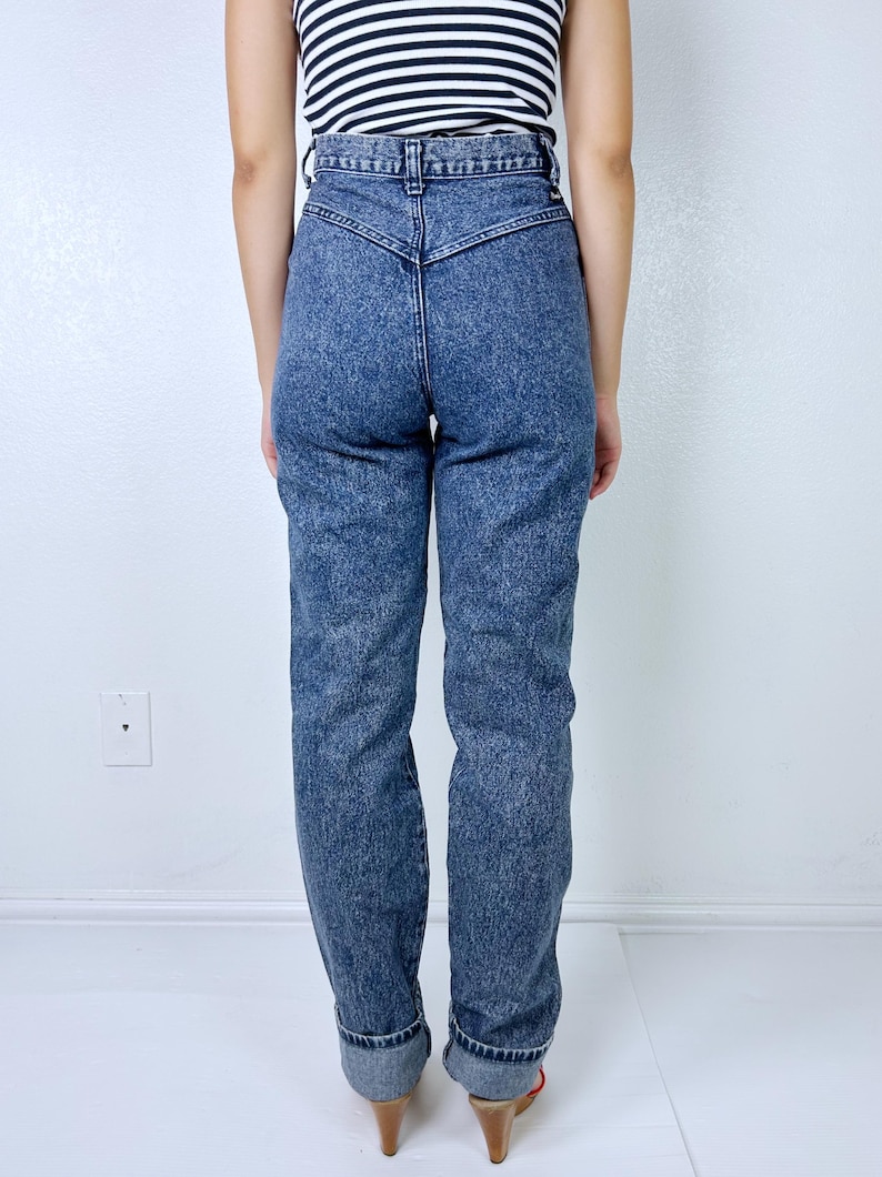 May include: Acid wash denim jeans with a high waist and tapered leg. The jeans have a classic five-pocket design and are cuffed at the ankles. The model is wearing a black and white striped top.