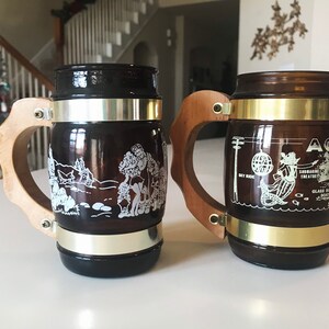 May include: Two brown glass beer steins with wooden handles and metal bands. One stein features a white graphic of a landscape, the other has a mermaid and text that reads "AQUA".