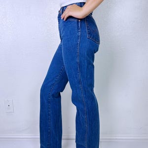 May include: High-waisted blue denim jeans featuring a straight-leg cut, a classic five-pocket design, and visible stitching along the side seams. The jeans have a medium-wash finish and a button-fly closure, shown from a side profile view.