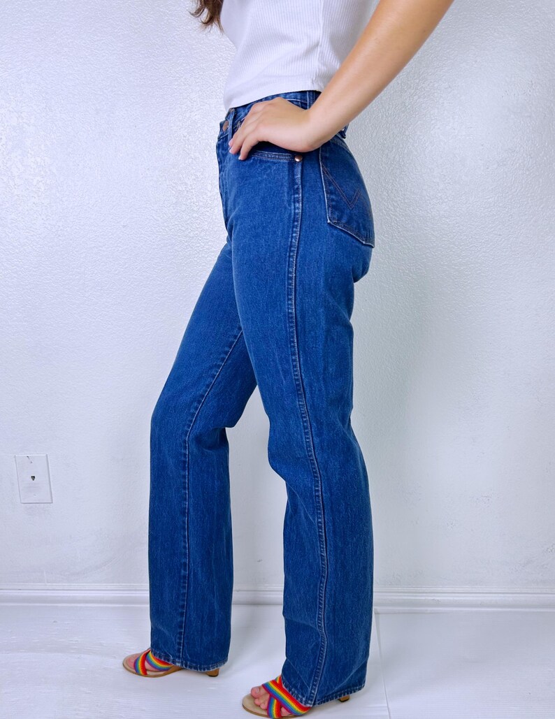 May include: High-waisted blue denim jeans featuring a straight-leg cut, a classic five-pocket design, and visible stitching along the side seams. The jeans have a medium-wash finish and a button-fly closure, shown from a side profile view.