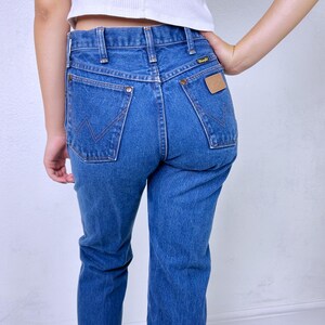 May include: Blue denim jeans featuring a high-waisted fit, a classic five-pocket design, and a tan leather patch on the back pocket. The back pockets are detailed with a signature W-shaped stitching pattern and copper-colored metal rivets.