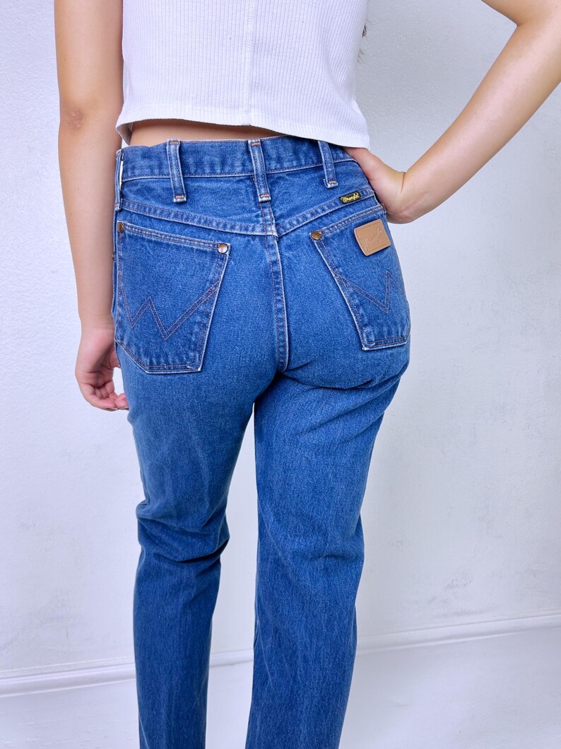 May include: Blue denim jeans featuring a high-waisted fit, a classic five-pocket design, and a tan leather patch on the back pocket. The back pockets are detailed with a signature W-shaped stitching pattern and copper-colored metal rivets.