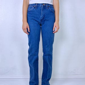 May include: A pair of high-waisted blue denim jeans with a straight-leg cut, featuring a classic five-pocket design, a metal button closure, and a zip fly. The jeans are paired with a plain white sleeveless top and rainbow-colored sandals.