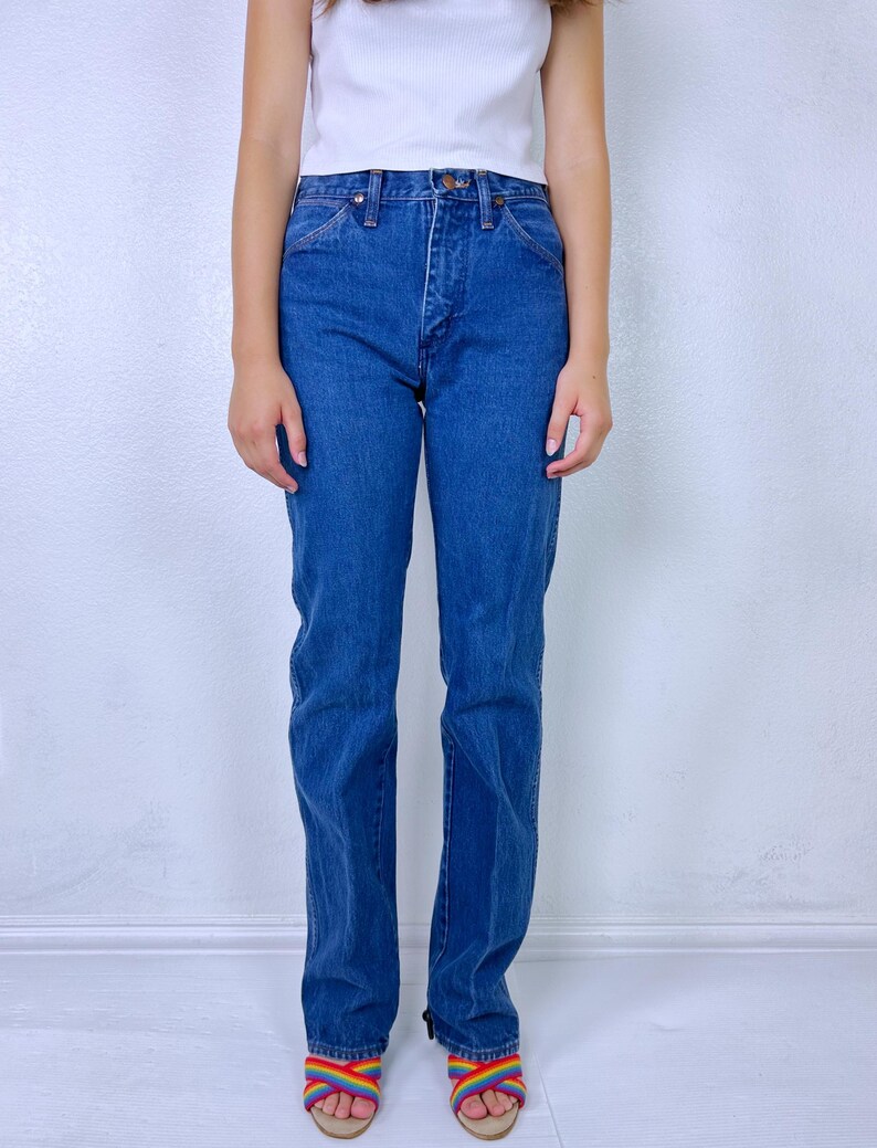May include: A pair of high-waisted blue denim jeans with a straight-leg cut, featuring a classic five-pocket design, a metal button closure, and a zip fly. The jeans are paired with a plain white sleeveless top and rainbow-colored sandals.
