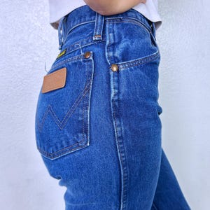 May include: High-waisted blue denim jeans featuring a tan leather patch on the back pocket with the Wrangler logo, copper-colored rivets, and a signature W-shaped stitching pattern on the pocket, paired with a white ribbed tank top.