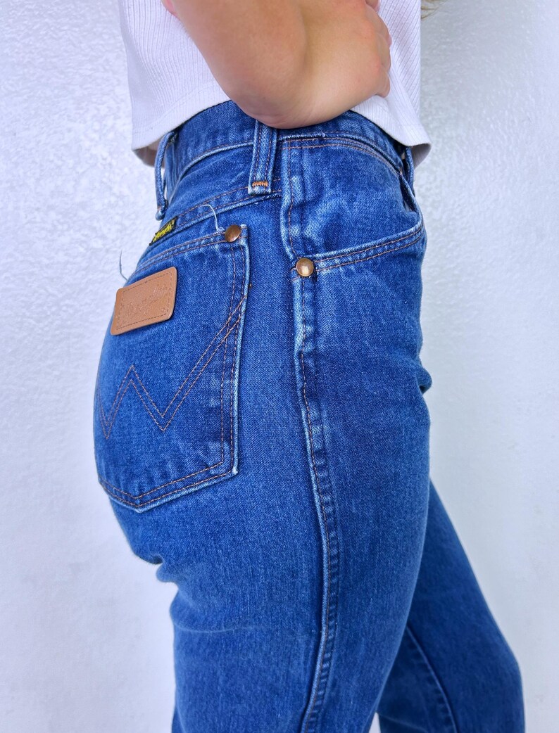 May include: High-waisted blue denim jeans featuring a tan leather patch on the back pocket with the Wrangler logo, copper-colored rivets, and a signature W-shaped stitching pattern on the pocket, paired with a white ribbed tank top.