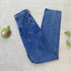 May include: Acid wash denim jeans with a heart-shaped design on the back. The jeans are a medium wash blue and have a classic five-pocket design. A green turtle and two green bunny figurines are on the side.
