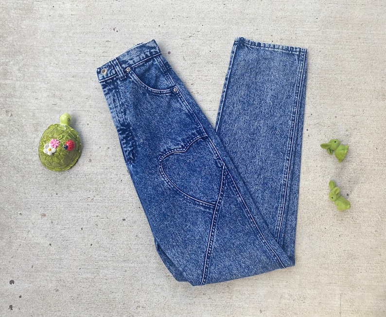 May include: Acid wash denim jeans with a heart-shaped design on the back. The jeans are a medium wash blue and have a classic five-pocket design. A green turtle and two green bunny figurines are on the side.