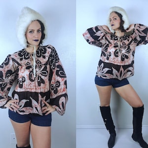 May include: A patterned blouse with long sleeves and a drawstring neckline. The blouse features a floral design in shades of brown, black, and red. The model is also wearing a white fur hat and denim shorts.