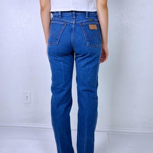 May include: A pair of medium-wash blue denim jeans featuring a high-waisted fit, straight legs, and two back pockets with a signature W-stitch design. A small tan leather patch is visible on the right back pocket with the Wrangler brand logo.