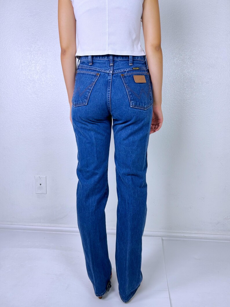 May include: A pair of medium-wash blue denim jeans featuring a high-waisted fit, straight legs, and two back pockets with a signature W-stitch design. A small tan leather patch is visible on the right back pocket with the Wrangler brand logo.