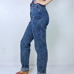 May include: Acid wash denim jeans with a high waist and a unique pocket design. The jeans are cuffed at the ankles. The person is wearing a black and white striped top and rainbow-colored sandals with a wooden heel.