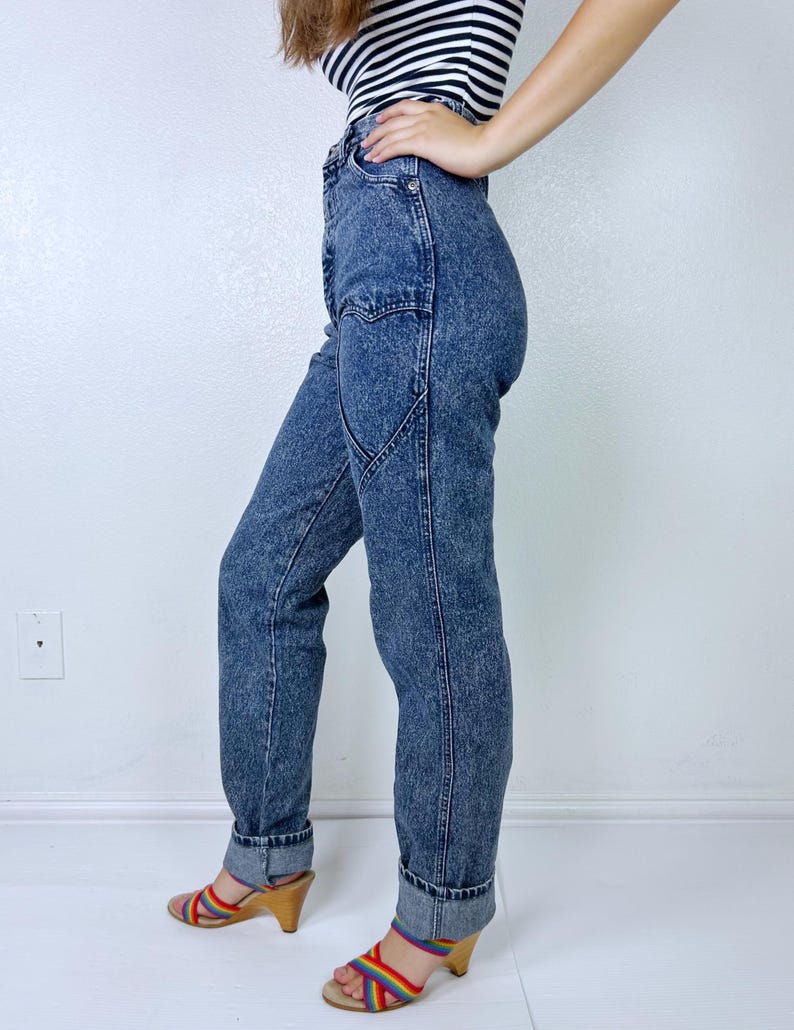 May include: Acid wash denim jeans with a high waist and a unique pocket design. The jeans are cuffed at the ankles. The person is wearing a black and white striped top and rainbow-colored sandals with a wooden heel.