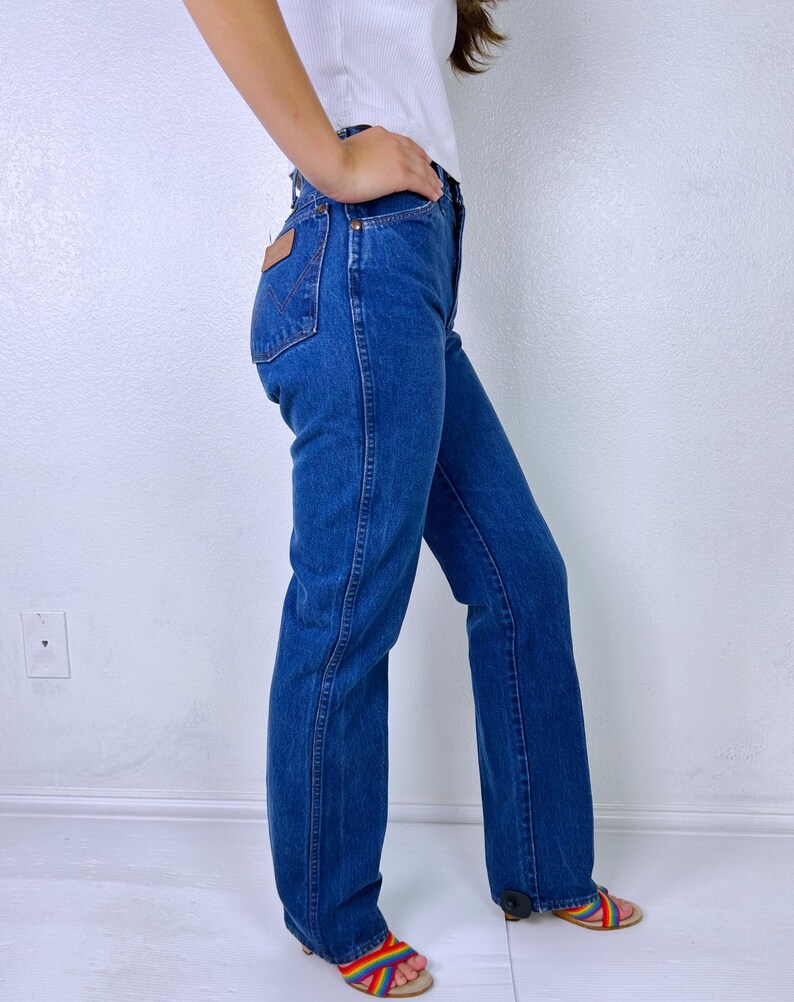 May include: High-waisted blue denim jeans featuring a straight-leg cut, a classic five-pocket design, and a tan leather patch on the back pocket with the Wrangler brand logo. The jeans are styled with a white top and rainbow-patterned sandals.