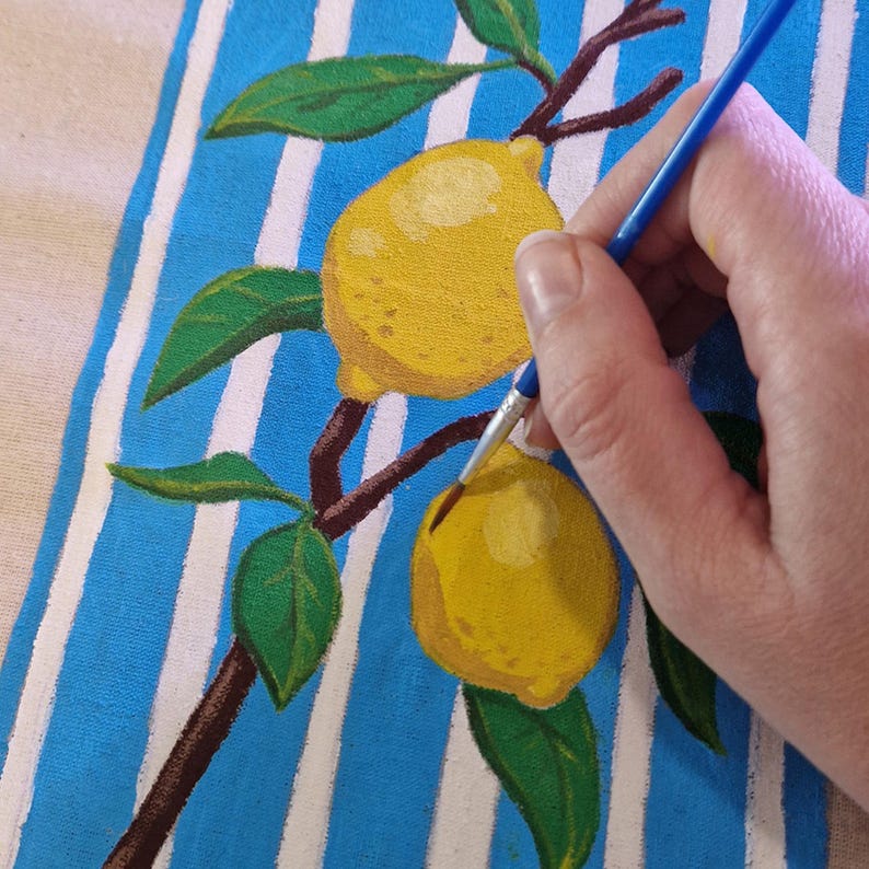 May include: A blue and white striped fabric surface featuring a hand-painted design of two yellow lemons on a brown branch with green leaves. A thin paintbrush with a blue handle is applying yellow paint to the lower lemon on the fabric.