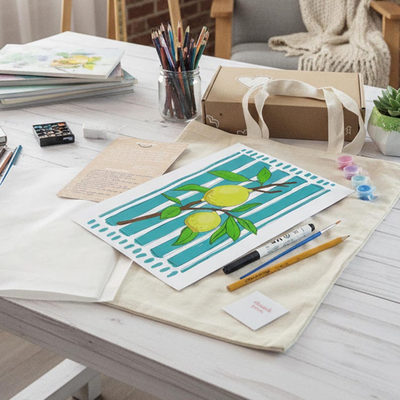 May include: A white canvas tote bag featuring a printed illustration of two yellow lemons on a branch with green leaves, set against a background of vertical teal stripes. The bag is displayed on a white wooden surface with art supplies.