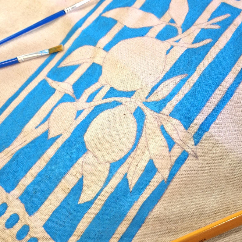 May include: A beige fabric surface featuring a hand-painted design of two lemons on a branch with leaves, set against a background of vertical bright blue stripes. Two thin paintbrushes with blue handles are positioned near the top left corner.