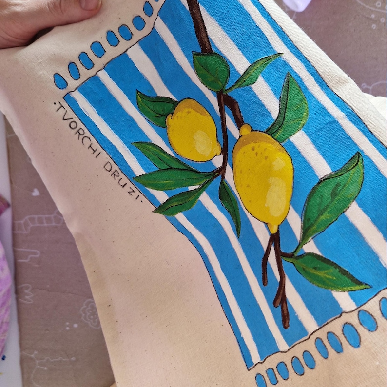 May include: A beige canvas tote bag featuring a hand-painted design of two yellow lemons on a brown branch with green leaves. The background has vertical blue and white stripes with blue oval accents at the top and bottom. Text reads TVORCHI DRUZI.